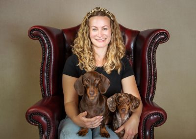 Lady sitting in a red Queen Ann chair with her 2 dachshund puppies on her lap